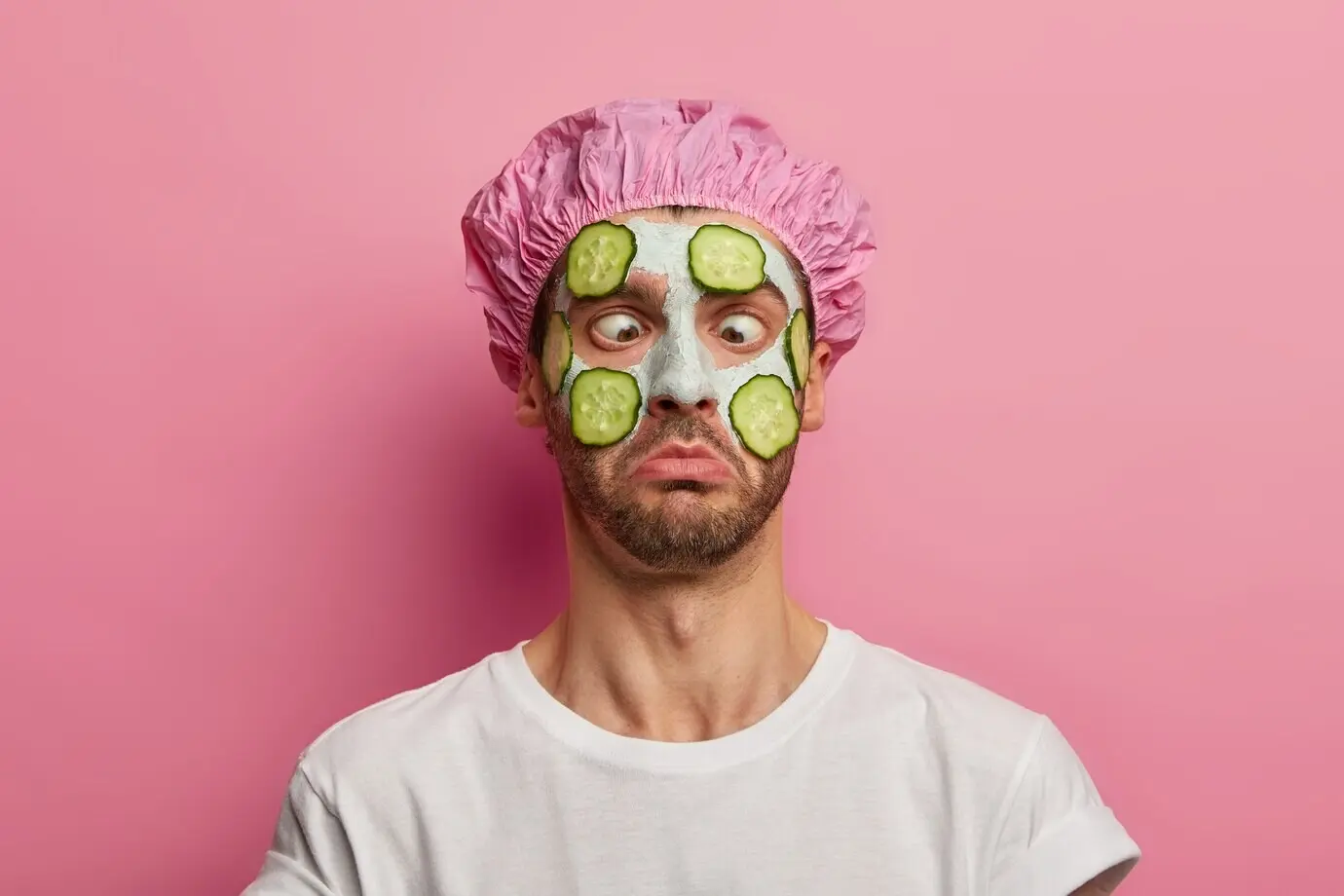 Молодой мужчина сводит глаза к переносице, наносит на лицо кремовую маску с огурцом, проводит бьюти-сеанс, в шапочке для душа.