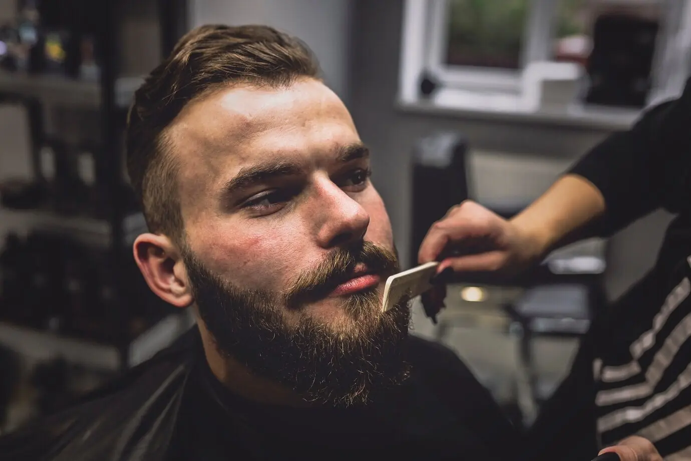 Обрезанный кадр: барбер расчёсывает бороду мужчине.